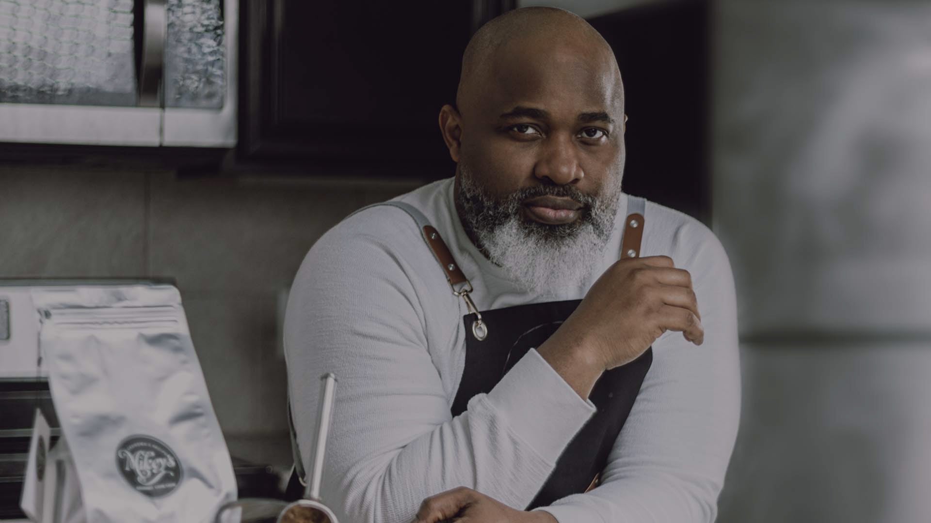 Michael Lawanson, the conservator of the family recipe for Mikey's Gourmet Chin-Chin, wearing a leather-strapped apron in a modern kitchen. A pack of Mikey's Gourmet Chin-Chin is displayed prominently in the foreground, emphasizing the brand's handcrafted quality and authentic recipe heritage.