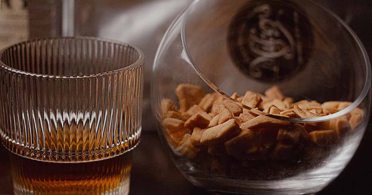 Mikey's Gourmet Chin-Chin in a glass bowl alongside a whisky, styled on a dark surface