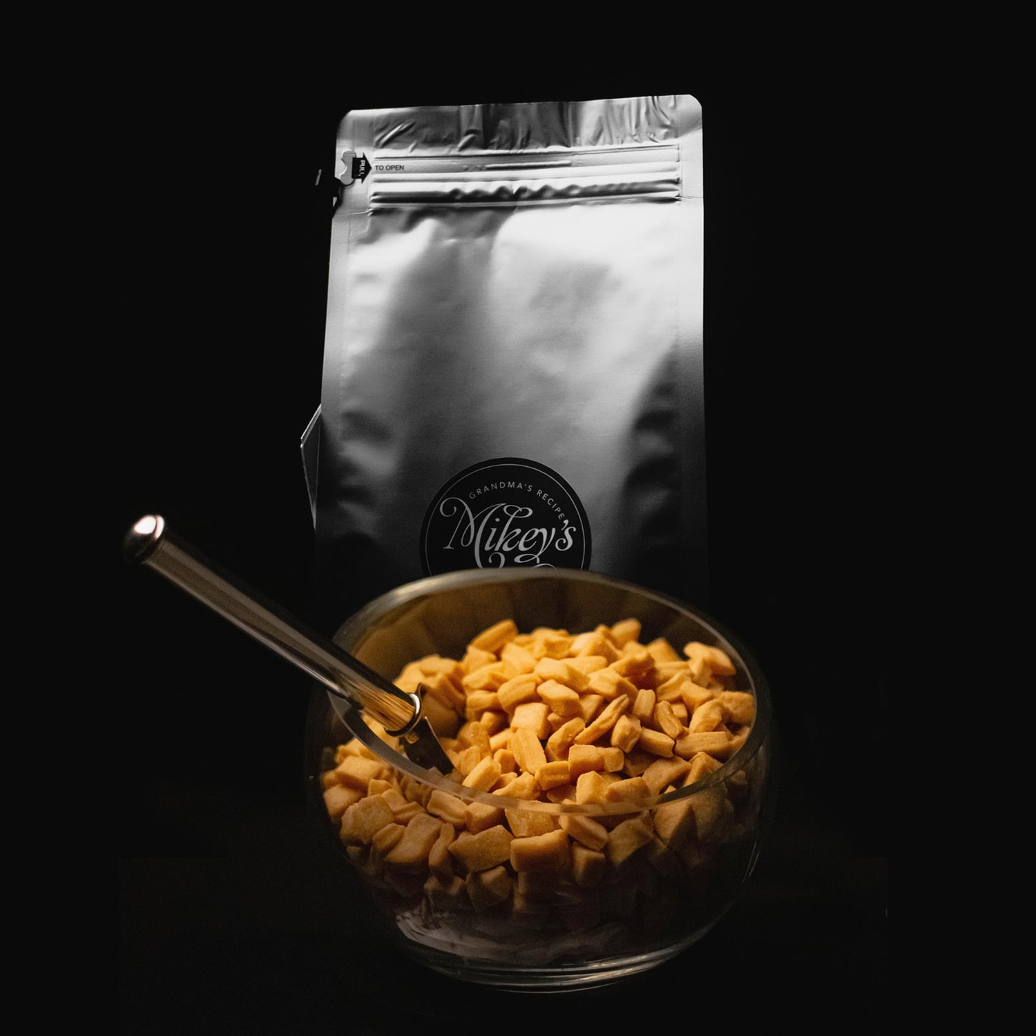 A bowl of Mikey's Gourmet Chin-Chin, a Nigerian-inspired snack, placed in front of sleek silver packaging featuring the Mikey's logo. The bowl is filled with golden, crunchy Chin-Chin pieces and accompanied by a silver spoon. Set against a black background, this image highlights the snack's authentic flavor and artisan presentation.