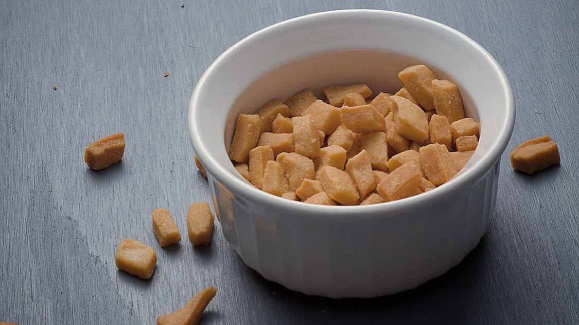 Mikey's Gourmet Chin-Chin golden pieces in a white ceramic bowl on a grey surface, with scattered pieces around it showing the crisp texture of this small-batch Nigerian snack.